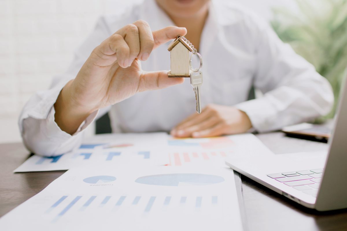 A person in a white shirt holds a small wooden house keychain with keys over financial documents, symbolizing home insurance and its importance