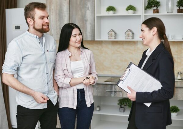 A smiling couple discusses home insurance with a professional agent holding a clipboard in a modern kitchen setting.